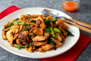 A plate of Pad See Ew with stir-fried rice noodles, chicken, and vegetables, served with a lime wedge.